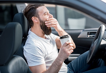 Man yawning with cup of coffee behind the wheel of a car